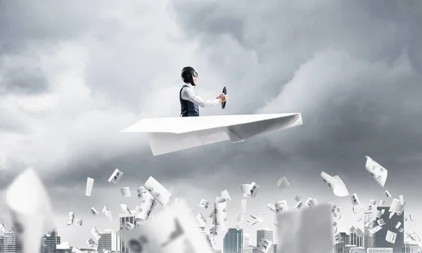 Crisis management and control in difficult situation concept. Businessman in aviator hat driving paper plane in storm. Pilot flying in small airplane. Megalopolis panorama with dramatic sky.