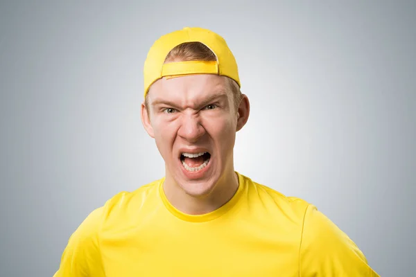 怒れる若者が叫んでいる 感情的な赤毛の少年は激怒した表情をしています 男の肖像画はグレーの背景に黄色のTシャツと野球帽を身に着けています 感情と表現の概念 — ストック写真