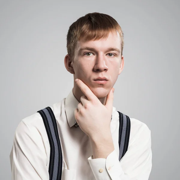Boy having serious and calm face with arms touching his chin. Caucasian redhead student has confident facial expression. Portrait of guy wears white shirt, bow tie and suspenders on grey background