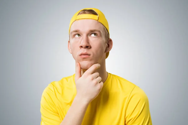 赤毛の生徒は上を向いて何かを思い出そうとします パズルしたティーンエイジャーは深刻な顔の表情をしています 男の肖像画は 灰色の背景に黄色のTシャツと野球帽を身に着けている — ストック写真
