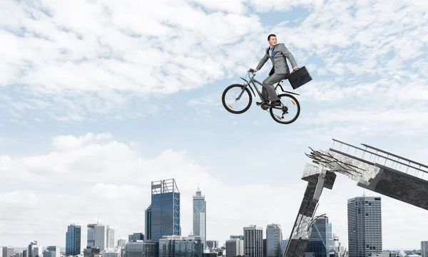 Man on bike jumping from broken bridge. Businessman looking back on bike and hurry to work. Employee in business suit with suitcase on cycle flying in air. Cyclist on background cityscape and blue sky