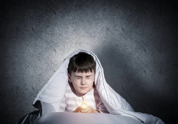 毛布の下に懐中電灯が隠れているおびえた子供 目を閉じた子供が家でベッドに横たわっている怖い子供 小さな男の子は夜眠れない 灰色の壁を背景にパジャマ姿の少年の肖像 — ストック写真
