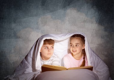 Dalmış küçük kız ve erkek yatmadan sonra yatakta kitap okuyor. Güzel çocuklar battaniyenin altında saklanıyorlar. Pijamalı okul öncesi çocuklar peri masalları okuyor. Gri duvar arka planlı yatak odası.