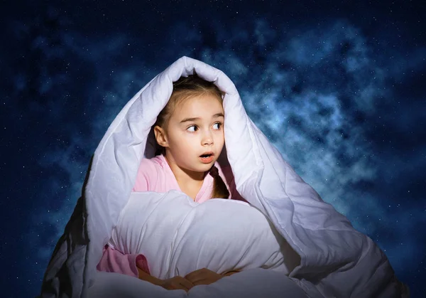 毛布の下に枕を隠して驚いた女の子 驚いた子供が自宅のベッドに横たわっていた 暗闇の恐怖覆われた子供は夜に眠ることができない 深い青空を背景にパジャマ姿の少女の肖像 — ストック写真