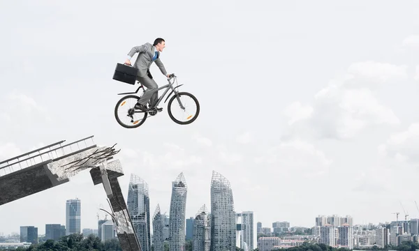 Man on bike jumping from broken bridge. Businessman on bike hurry to work. Corporate employee in business suit with suitcase on cycle flying in air. Cyclist on background modern cityscape and blue sky