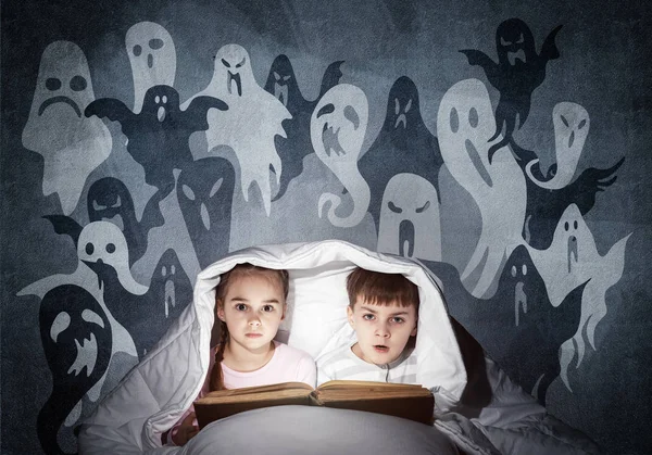 害怕的女孩和男孩在床上读孩子的书 惊慌失措的孩子一起躺在毯子下 穿着睡衣和滑稽鬼的孩子在灰墙上剪影 儿童阅读魔法童话 — 图库照片