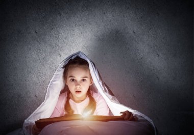 Yatmadan önce yatakta kitap okuyan korkmuş küçük kız. Battaniyenin altında el feneri saklanan güzel bir çocuk. Çocuk geceleri uyuyamıyor. Gri duvarın arka planında pijamalı kız portresi.