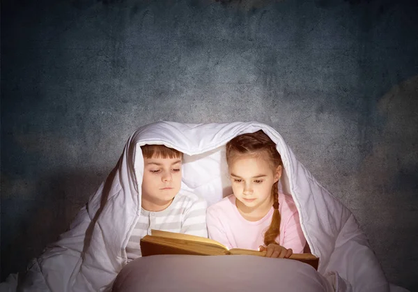 小さな女の子と男の子が毛布の下でお互いを見ている 寝る前にベッドで魔法の物語を読む子供たち 灰色の壁を背景にパジャマの妹と兄弟 — ストック写真