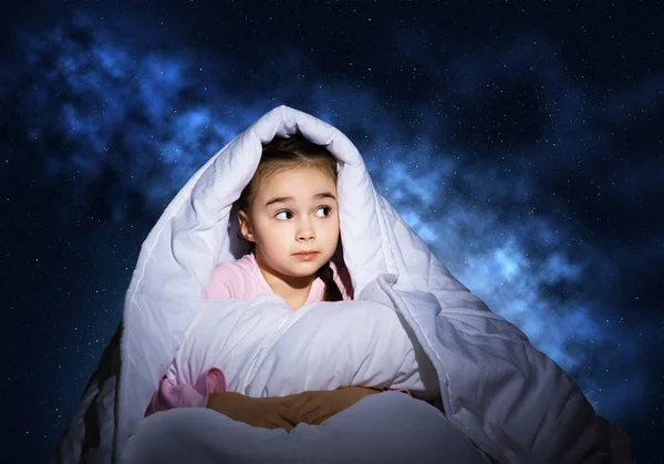 Scared girl with pillow hiding under blanket. Startled kid lying in his bed at home. Little girl afraid of dark. Covered child not sleep at night. Girl in pajamas on background of deep blue sky.