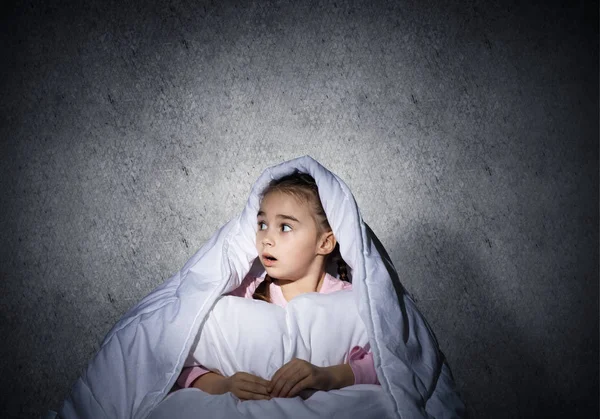 Frightened girl hiding under blanket. Scared kid lying in his bed at home. Fear of the dark. Beautiful little girl can not sleep at night. Portrait of child in pajamas on background of grey wall.
