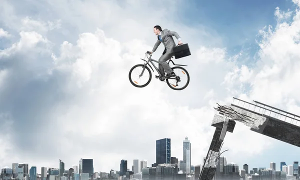 Man on bike jumping from broken bridge. Businessman on bike hurry to work. Corporate employee in business suit with suitcase on cycle flying in air. Cyclist on background modern cityscape and blue sky