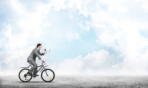 Businessman with megaphone on bike at sunny day. Marketing and advertising campaign. Manager in business suit riding bicycle and speaking in loudspeaker. Male cyclist on background of blue sky.