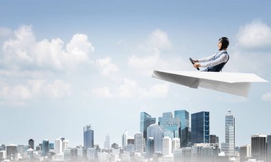 Havacı şapkalı işadamı kağıt düzlemde oturuyor ve direksiyonu tutuyor. Pilot yüksek gökdelenler ve ofis binaları ile iş merkezi üzerinde kağıt uçak sürüş. Bulutlu mavi gökyüzü arka plan