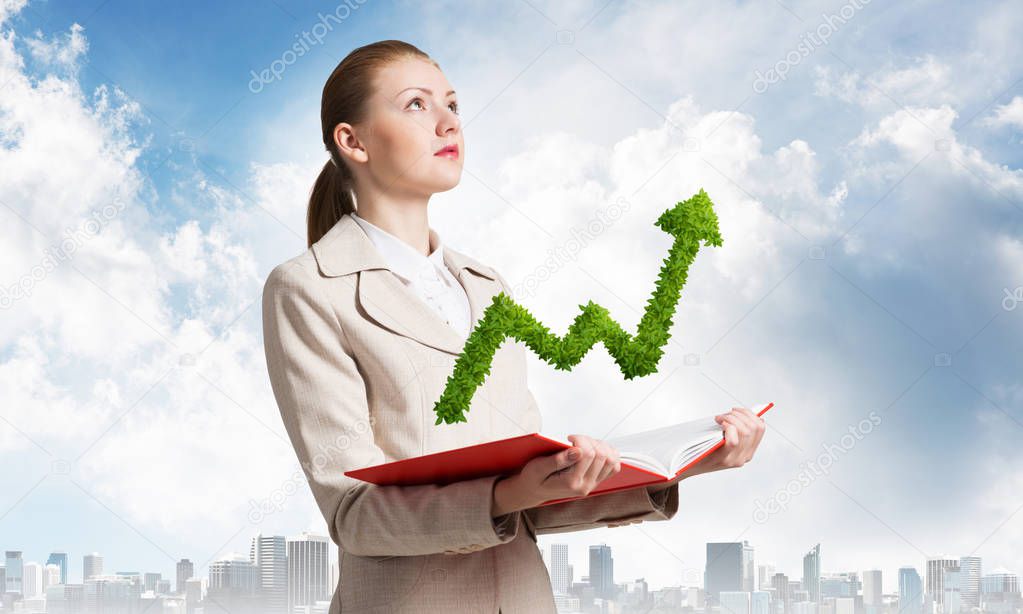Businesswoman with green plant shaped growing arrow graph above opened notebook. Business statistics and analytics. Elegant young woman in white business suit on background of cityscape and blue sky.