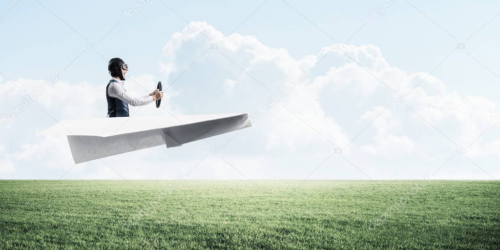 Happy aviator driving small paper plane on background of natural landscape. Man in big paper airplane flying low above green meadow. Nature panorama with green grass and cloudy sky in summer day.