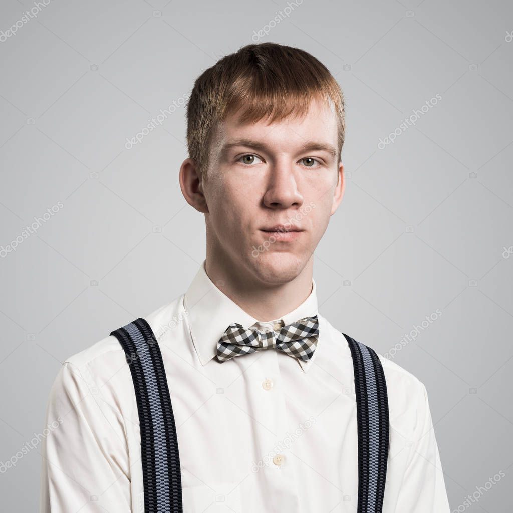 Boy having serious and calm face. Caucasian redhead student has confident facial expression. Portrait of guy wears white shirt, bow tie and suspenders on grey background. Emotion and expression