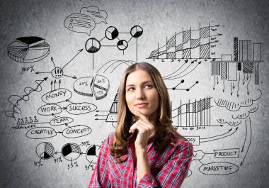 Kahverengi saçlı kadın dalgın görünüyor ve bir şeyler düşünüyor. Finansal tablolar ve gri duvara iş karalamaları. Kafası karışmış kızın ciddi bir yüz ifadesi var. Kadın kırmızı kareli gömlek giyiyor.
