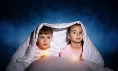 Yatakta el feneri olan çocuklar. Küçük kız ve erkek kardeş battaniyenin altında saklanıyorlar. Derin mavi gökyüzünün arka planında geceleri karanlıktan korkan kapalı çocuklar. Çocuğun gece terörü.