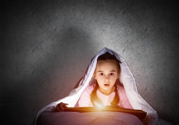 寝る前にベッドで本を読んで怖がっていた少女 毛布の下に隠れて懐中電灯を持つかなりの子供 子供の夜に眠ることができない 灰色の壁を背景にパジャマ姿の少女の肖像 — ストック写真