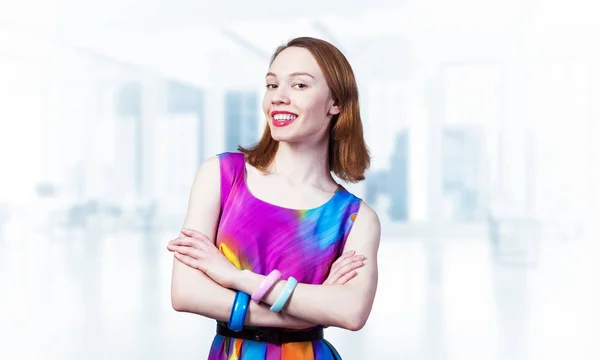 Happy woman standing with folded arms. Elegant good looking lady wears bright dress and bracelets. Joyful redhead girl with perfect teeth smile on blurred office background. Flirty student posing