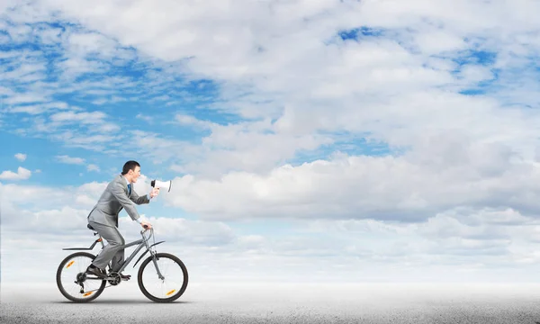 Businessman with megaphone on bike at sunny day. Marketing and advertising campaign. Manager in business suit riding bicycle and speaking in loudspeaker. Male cyclist on background of blue sky.