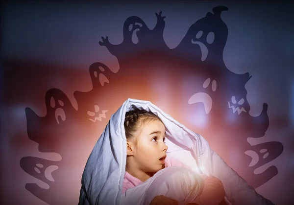 Excited girl hiding under blanket from imaginary ghosts. Scared kid sitting in his bed at home. Little girl afraid of fantasy monsters. Covered child in pajamas not sleep at night. Halloween holiday.
