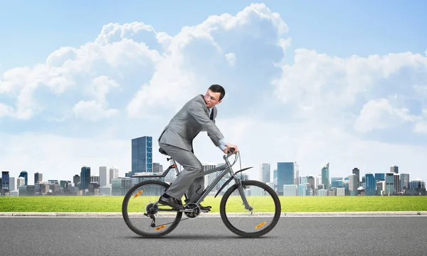 Businessman commuting to work by bike. Man wearing business suit riding bicycle on asphalt road. Handsome cyclist looking back on background of modern cityscape and blue cloudy sky.