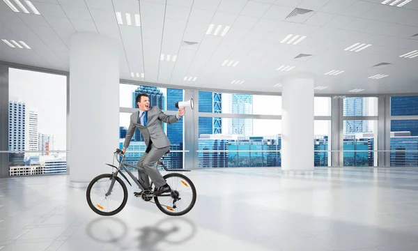 会議室で自転車に乗るビジネススーツの男 パノラマの窓のあるロフトオフィスのインテリアで自転車を振り返ってメガホンを持つビジネスマン 事業説明会及び発表 — ストック写真