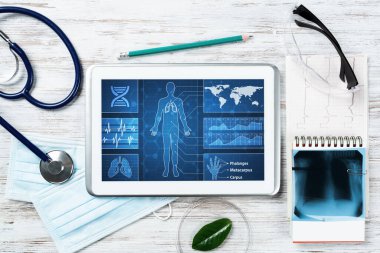 Doktorların ofisinde laboratuvar hasta muayenesi. Ekranda tıbbi uygulama arayüzü olan tablet bilgisayar. Stetoskop, röntgen görüntüsü ve kardiyogram tahta masada. Tıbbi teşhis ve muayene