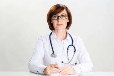 Ofiste oturan çekici kızıl saçlı doktor kameraya bakıyor. Beyaz önlüklü, steteskoplu beyaz arka planda duran terapist portresi. Profesyonel tıbbi teşhis ve tedavi.