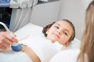 Doktor ultrason hastanedeki küçük hastanın karnını muayene ediyor. Modern ekipmanlarla klinikte karın boşluğu teşhisi konuldu. Yakın plan mutlu küçük kız yatakta yatıyor. En kaliteli çocuk sağlık hizmetleri.