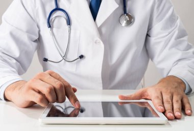 Erkek terapist ellerini tablet bilgisayar kullanarak kapat. Masasında steteskoplu beyaz üniformalı bir doktor oturuyor. İnceleme ve tanı konularında modern teknoloji. Tıbbi dijital uygulama