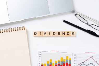 Tahta küplerdeki harflerle ödeme konseptini böler. Malzemelerle dolu ofis hayatı. Düz beyaz yüzey dizüstü bilgisayarı, not defteri ve mali raporuyla yatıyordu. Sermaye tasarrufu ve yatırım