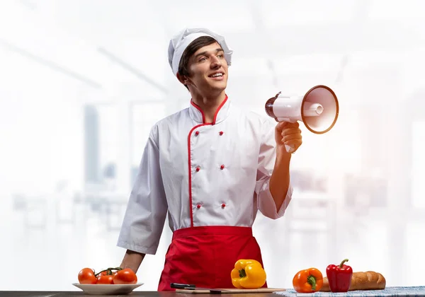 年轻的厨师手里拿着扩音器站着 情绪激动 头戴白帽 头戴红围裙 头戴浅色厨房内饰的高加索厨师 餐厅广告 食品零售活动公告 饮食服务 — 图库照片