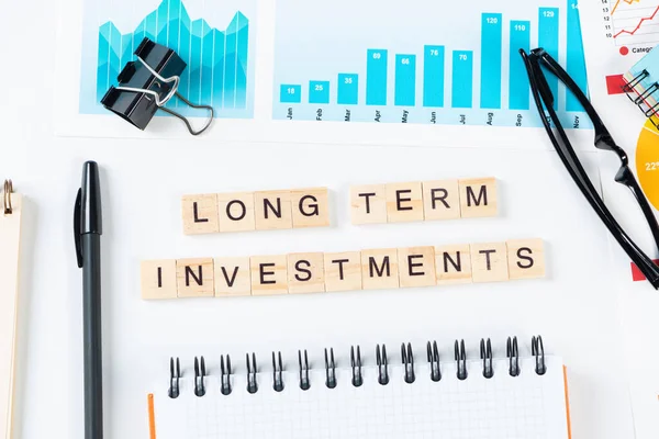 Long term investments concept with letters on wooden cubes. Still life of office workplace with supplies. Flat lay white surface with notepad, pen and financial report. Financial success and planning