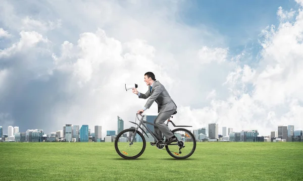 Businessman with megaphone on bike at sunny day. Marketing and advertising campaign. Manager in business suit riding bicycle on green meadow. Male cyclist on background of blue sky and city on horizon