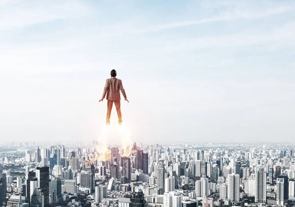 Businessman in suit and aviator hat flying in blue sky as superhero. Corporate manager as superman launching upwards with jet flame above modern city. Successful business startup motivation concept