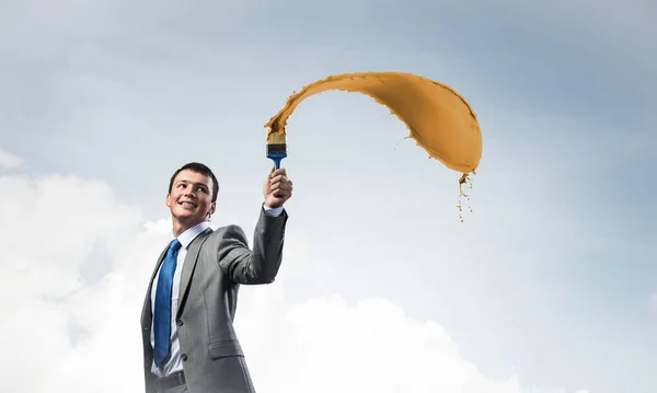 Smiling creative businessman painter holding paintbrush. Portrait of joyful handsome man in business suit and tie. Orange paint splash on blue sky background. Ambitions and creativity in business