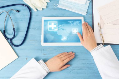 Tablet bilgisayarlı doktor elleri. Mavi ahşap masada oturan beyaz önlüklü bir terapist. Klinikte muayene ve konsültasyon. Tıbbi başvuru ve çevrimiçi sağlık hizmetleri.