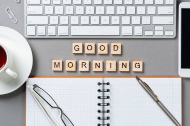 Tahta küplerdeki harflerle günaydın mesajı. Malzemelerle dolu çalışma alanı. Düz gri yüzey, bilgisayar klavyesi ve spiral not defteriyle yatıyordu. Sabah tebrikleri ve motivasyon.