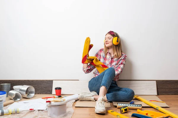 快乐的女孩 戴着防护耳机和手套坐在地板上 搬家后的家庭改造 建筑工具和建筑材料 穿着格子衬衫和牛仔裤的年轻女子 — 图库照片