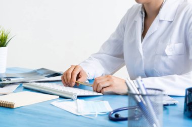 Kadın doktor elinin kalemle yakın çekimi. Masada bilgisayarla çalışan tıp üniformalı bir doktor. İnceleme ve tanı konularında modern teknoloji. Bilgisayar klavyeli pratik işyeri