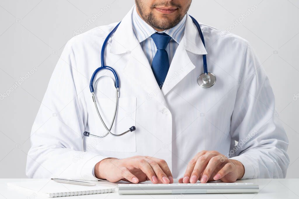 Male therapist typing at wireless computer keyboard. Physician in medical uniform with stethoscope working with computer at desk. Modern technology in examination and diagnosis. Health care concept