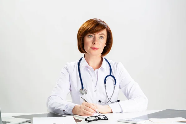 オフィスで紙に書いている美しい赤毛の医者 デスクに座って聴診器を備えた白いコートの医師 診療所での専門的な医療診断と治療 — ストック写真
