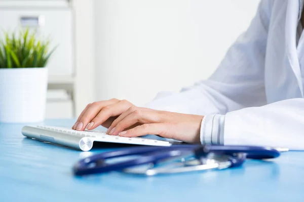 Doctor typing at computer keyboard. Physician in medical uniform working with computer at desk in office. Modern technology in examination and diagnosis. Doctor workplace with green plant in pot.