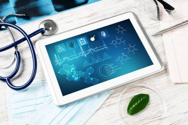 Doktorların ofisinde laboratuvar hasta muayenesi. Ekranda tıbbi uygulama arayüzü olan tablet bilgisayar. Stetoskop, röntgen görüntüsü ve kardiyogram tahta masada. Tıbbi teşhis ve muayene
