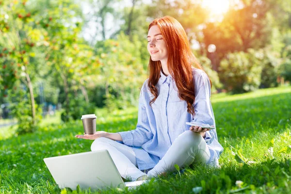 若い赤毛の女性は緑の芝生の上でノートパソコンとコーヒーカップで蓮のポーズで瞑想します 閉じた目でヨガの練習カジュアルウェアの美しい女の子 夏の日の屋外の仕事とリラクゼーション — ストック写真