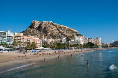 Alicante plajı boyunca insanların yaz güneşinin tadını çıkardığı panoramik bir manzara ve ünlü simgesi olan dağ sırtındaki şato.