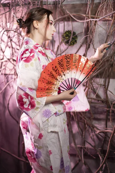 beautiful fair-skinned girl with Japanese makeup and Japanese clothes standing with a red fan near the tree branches and window