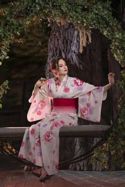 一个美丽的皮肤黝黑的女孩 日本化妆和日本服装坐在秋千上 扭曲的绿叶 独自在夜间 — 图库照片